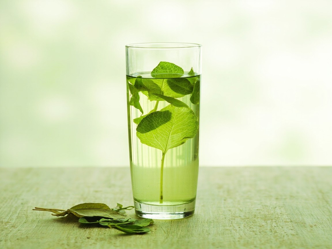 A tall clear glass tumbler filled with pale yellow-green tea liquor, with several green tea leaves visibly unfurled and suspended in the clear liquid, soft natural daylight backlighting the leaves through the glass